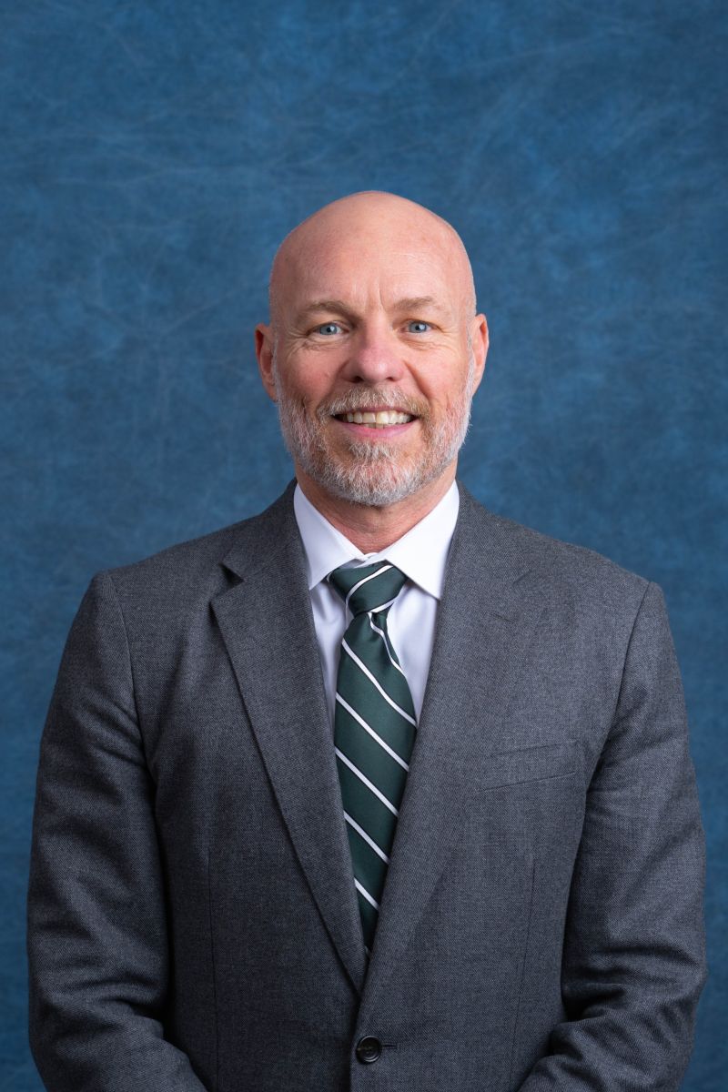 Dr. William Wells poses for a photo. Wearing a button down shirt. Smiling in front of a neon sign glowing Justice.